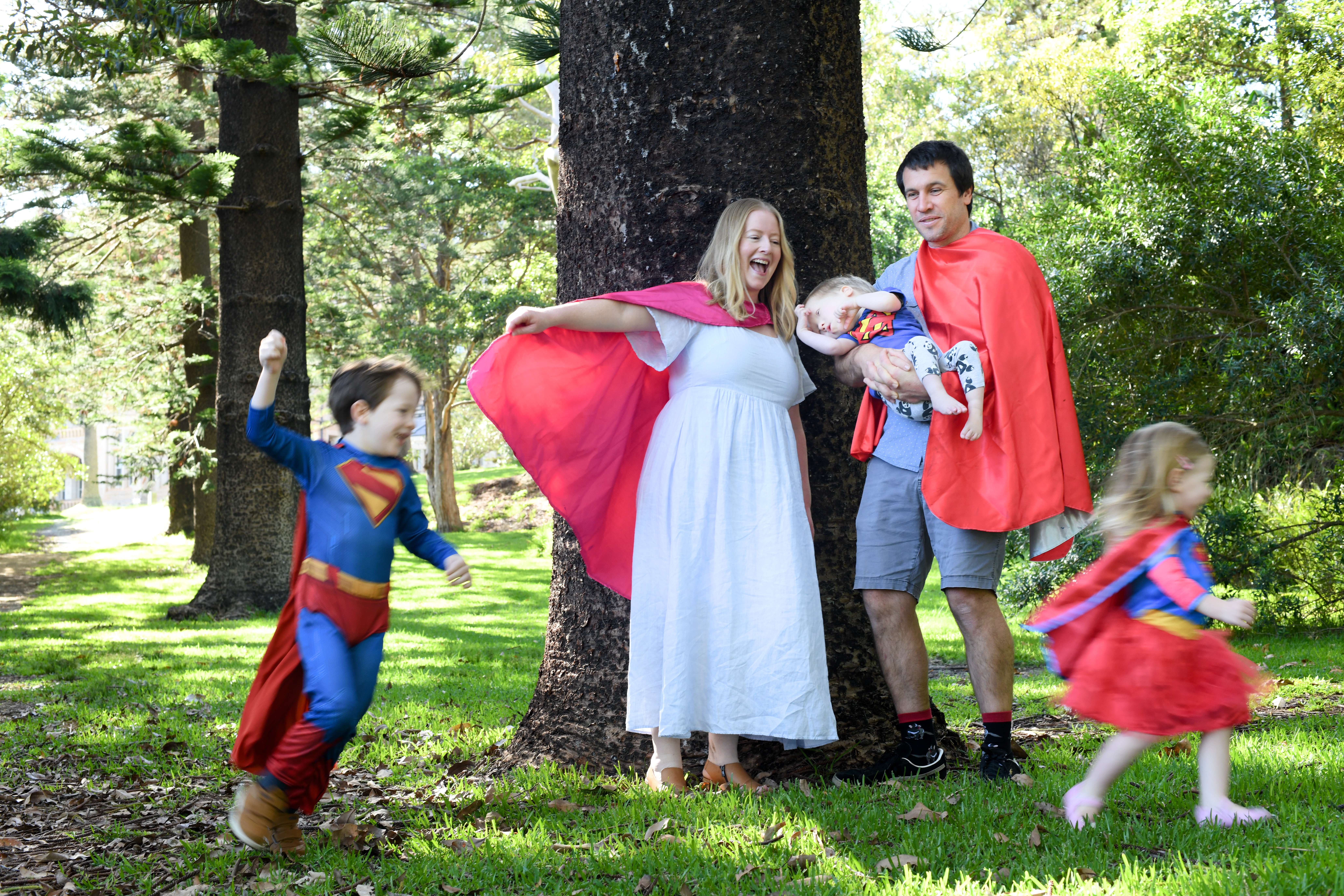 Mum and dad in superhero capes with two small children running in front of them. Dad is holding a young child.