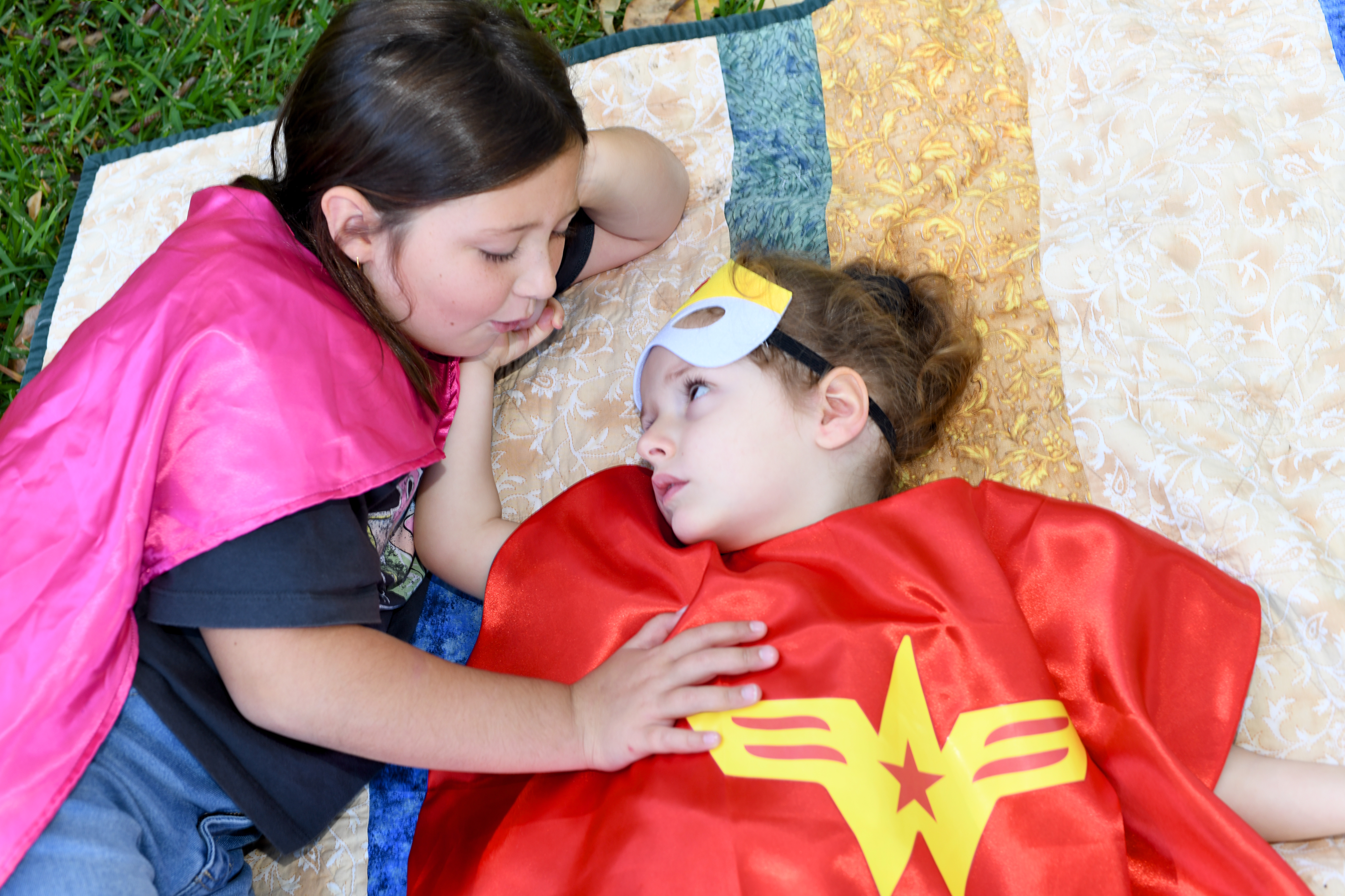 Bonnie laying on a blanket on the grass wearing a Wonderwoman cape with her sister laying beside her